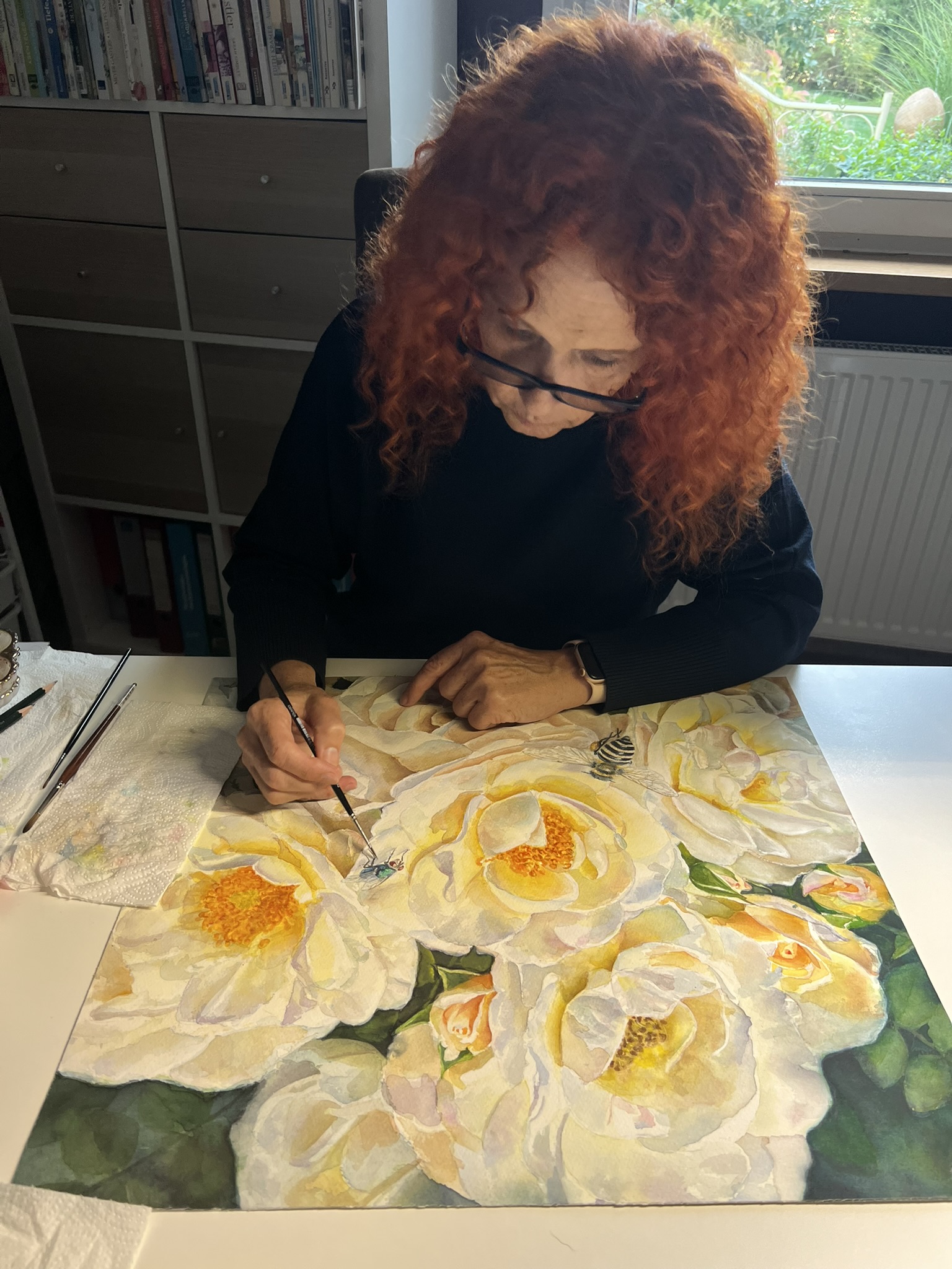Doris Joa painting the white rose watercolor with a bee and a fly in her studio, viewed from the front